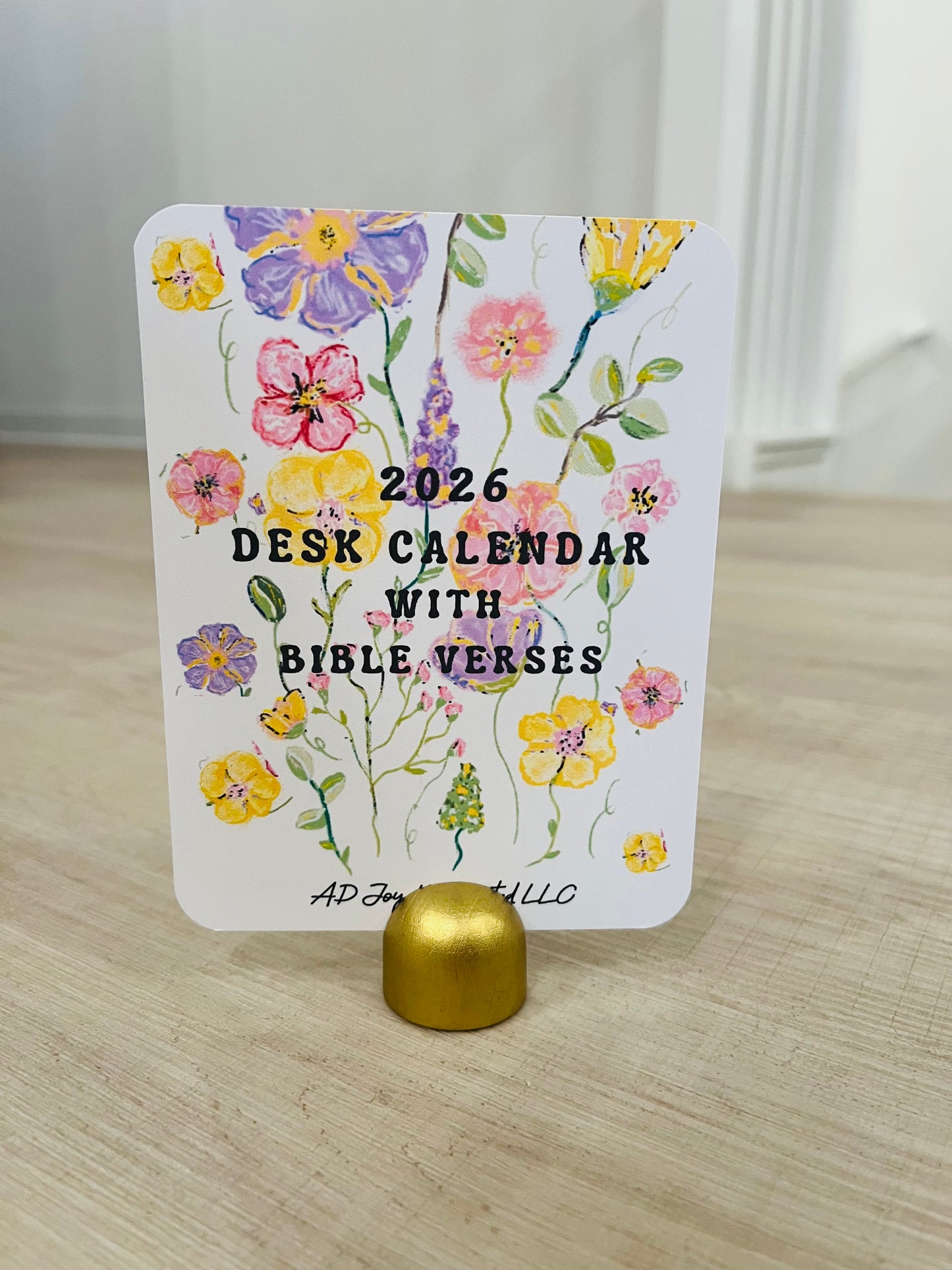 A packaged handmade 2025 calendar with one wood holder, featuring colorful cartoon animals and designs on the front, displayed on a table with green leaves in the background.