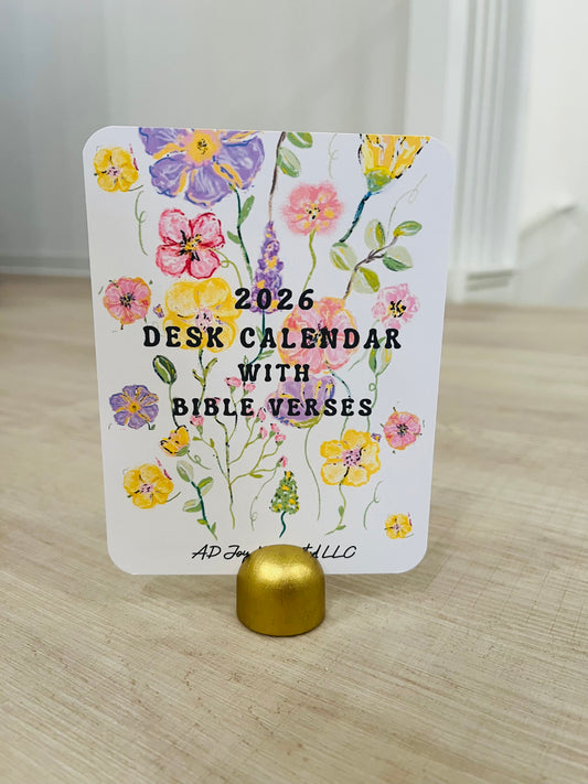 A packaged handmade 2025 calendar with one wood holder, featuring colorful cartoon animals and designs on the front, displayed on a table with green leaves in the background.